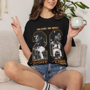 May include: A black short-sleeve t-shirt featuring a graphic print with the text TWO ICONS. ONE IMPACT. The design includes two large hexagonal frames containing black and white portraits of Usher and Chris Brown, with their names printed below.