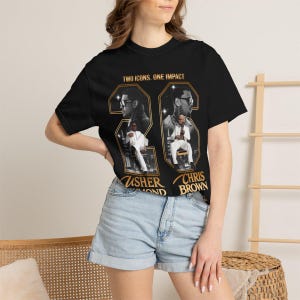 May include: A black short-sleeve t-shirt featuring a graphic print with the text "TWO ICONS. ONE IMPACT" at the top. The design includes two framed portraits of performers, with the names "USHER RAYMOND" and "CHRIS BROWN" printed in gold below.