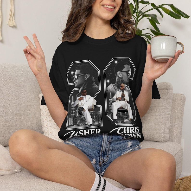 May include: A black short-sleeve t-shirt featuring a large graphic print of Usher and Chris Brown. The design includes the number 26, black and white portraits of the artists, and images of them in white suits against a city skyline background.