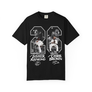 May include: A black short-sleeve t-shirt featuring a graphic print of Usher Raymond and Chris Brown. The design includes large numbers, city skylines, and the names Usher Raymond and Chris Brown in white text, along with their printed signatures below.