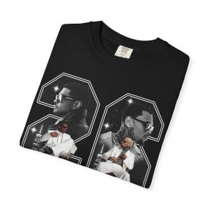 May include: A black t-shirt featuring a large graphic print of the number 24. Inside the digits are black and white photographic portraits of a person wearing sunglasses and a white suit, set against a starry night sky and city skyline background.