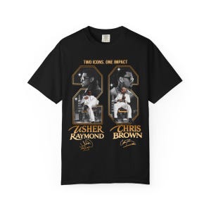 May include: A black short-sleeve t-shirt featuring a graphic print with the text "TWO ICONS. ONE IMPACT" above two framed portraits of Usher Raymond and Chris Brown. Each portrait includes the artist's name and a signature in gold lettering.