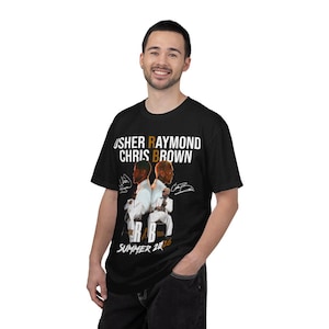 May include: A black t-shirt featuring a graphic print of Usher Raymond and Chris Brown in white suits. The text reads USHER RAYMOND CHRIS BROWN, THE R&B TOUR, and SUMMER 2026, accompanied by two signatures on either side of the central figures.