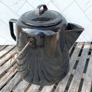 May include: Black enamel camping coffee pot with a lid and a spout. The pot has a speckled finish and a metal handle.
