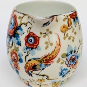 May include: A cream-colored ceramic creamer featuring a colorful floral pattern with red, blue, and yellow flowers interspersed with green vines and a prominent bird illustration with orange and yellow plumage perched on a branch.