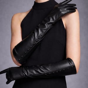 Peut inclure: Longues gants d'opéra en cuir noir avec des boutons. Les gants s'étendent au-delà des coudes et sont portés avec une robe noire. Design élégant, idéal pour les événements formels ou pour ajouter une touche de sophistication.