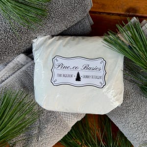 May include: A package of laundry detergent from Pine.co Basics, with the text "Four Ingredient - Laundry Detergent", rests on folded gray towels. Green pine needles are arranged around the package and towels, creating a natural aesthetic.