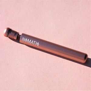 May include: A rose gold metal tube with a screw-on cap and the word "NOMATIQ" printed on the side. The tube is cylindrical and appears to be empty. The cap has a ridged design for easy grip.