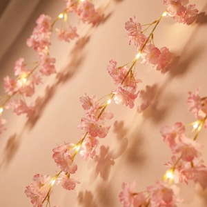Op de afbeelding: Slinger met roze kersenbloesemlichtjes. De lichten zijn kleine, warmwitte LED's, afgewisseld met delicate roze bloemen. De bloesems hebben een zachte roze kleur en het totale effect is romantisch en delicaat.
