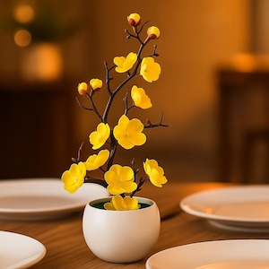May include: A white ceramic pot displays a decorative floral arrangement. The arrangement features a dark brown branch with bright yellow flowers and buds. The pot sits on a wooden table, with white plates in the background.