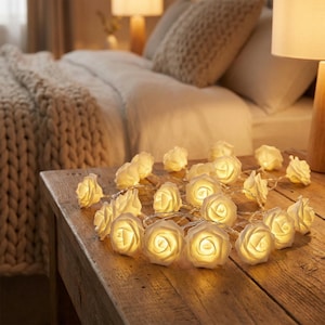 Può includere: Una stringa di luci a forma di rosa bianca su un tavolo di legno. Le luci illuminate emettono un bagliore caldo. Lo sfondo include un letto con una coperta di maglia beige e cuscini, suggerendo un'atmosfera accogliente.