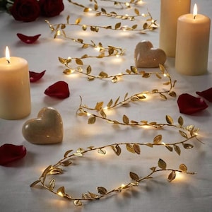 Pode incluir: Uma cena romântica com luzes de corda douradas em forma de folhas, velas e decorações em forma de coração. Pétalas de rosa vermelhas e rosas espalhadas. A luz quente das velas e das luzes cria um ambiente acolhedor, ideal para decoração de casa ou eventos especiais.