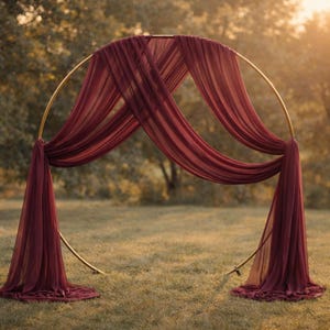 May include: A circular gold metal wedding arch adorned with draped burgundy fabric. The arch is positioned on grass, with the fabric arranged to create a decorative backdrop for weddings or other special occasions.