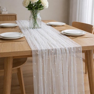 May include: A white, sheer table runner with pearl embellishments is laid across a wooden dining table. White plates and a vase of white flowers are also on the table. The runner adds a touch of elegance to the dining setting.