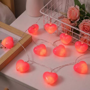 Könnte beinhalten: Eine Lichterkette mit herzförmigen Lichtern. Jedes Herz ist rosa und hat ein warmes, leuchtendes Zentrum. Die Lichter sind an einem dünnen, silbernen Draht befestigt und schaffen eine romantische und festliche Atmosphäre. Ideal für Valentinstag.