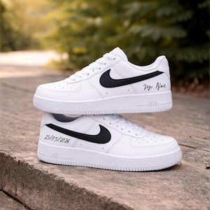 May include: Two white Nike Air Force 1 sneakers with black swooshes. One shoe has "Mr. Alex" written on the side, and the other has "23/05/2026". The shoes are stacked on top of each other.