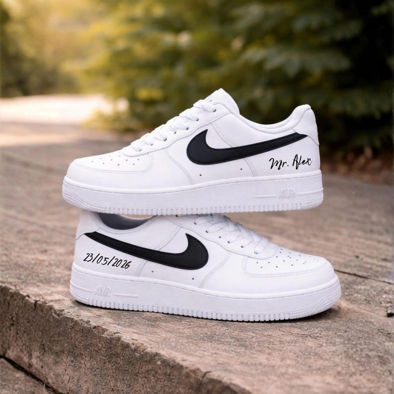 May include: Two white Nike Air Force 1 sneakers with black swooshes. One shoe has "Mr. Alex" written on the side, and the other has "23/05/2026". The shoes are stacked on top of each other.