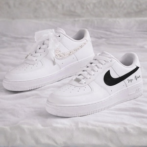 May include: Pair of white sneakers. One shoe features a pearl-studded swoosh and ribbon laces. The other shoe has a black swoosh and the name "Mr. Alex" in black script. The shoes are on a white surface.