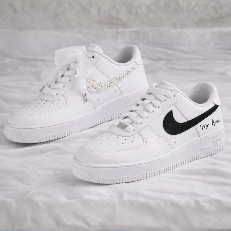 May include: Pair of white sneakers. One shoe features a pearl-studded swoosh and ribbon laces. The other shoe has a black swoosh and the name "Mr. Alex" in black script. The shoes are on a white surface.
