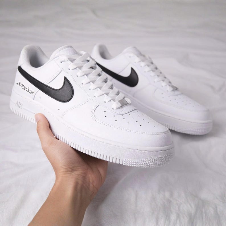 May include: A pair of white Nike Air Force 1 sneakers with black swoosh logos. The sneakers have white laces and soles. The date 23/05/2026 is printed on the side of one shoe.