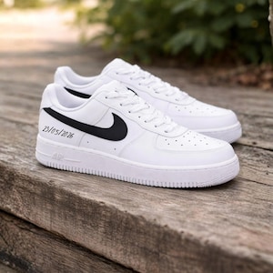 May include: Pair of white sneakers with black swoosh logos and laces. The shoes have the text "23/05/2026" on the side. The sneakers are on a wooden surface.