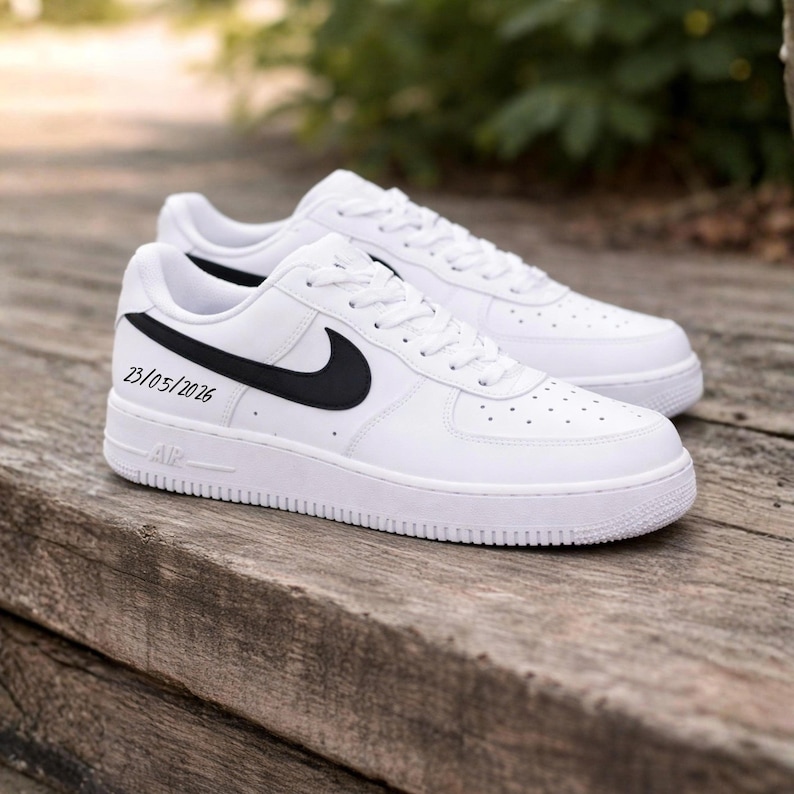 May include: Pair of white sneakers with black swoosh logos and laces. The shoes have the text "23/05/2026" on the side. The sneakers are on a wooden surface.