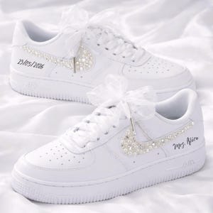 May include: White sneakers with pearl and rhinestone embellishments. The Nike swoosh is adorned with pearls. The laces are tied with white ribbons. One shoe has the date 23/05/2026, and the other has the text "Mrs. Alice".