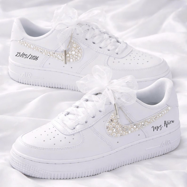 May include: White sneakers with pearl and rhinestone embellishments. The Nike swoosh is adorned with pearls. The laces are tied with white ribbons. One shoe has the date 23/05/2026, and the other has the text "Mrs. Alice".
