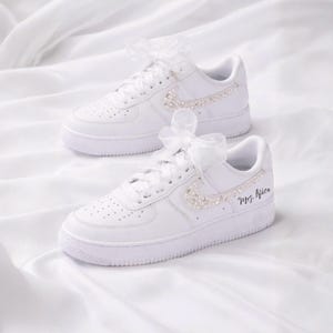 May include: White sneakers with pearl and rhinestone embellishments on the Nike swoosh. The laces are tied with white ribbons. The right shoe has the text "Mrs. Alice" in black script.
