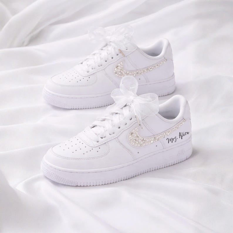 May include: White sneakers with pearl and rhinestone embellishments on the Nike swoosh. The laces are tied with white ribbons. The right shoe has the text "Mrs. Alice" in black script.