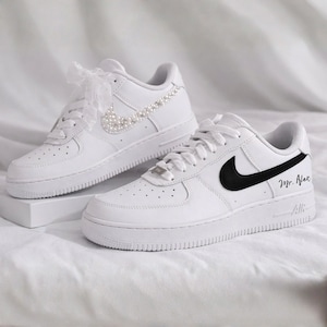May include: A pair of white trainers with black swooshes. One trainer is decorated with pearls and a sheer ribbon. The other has the text "Mr. Alex" in black script. Both have white laces and soles.