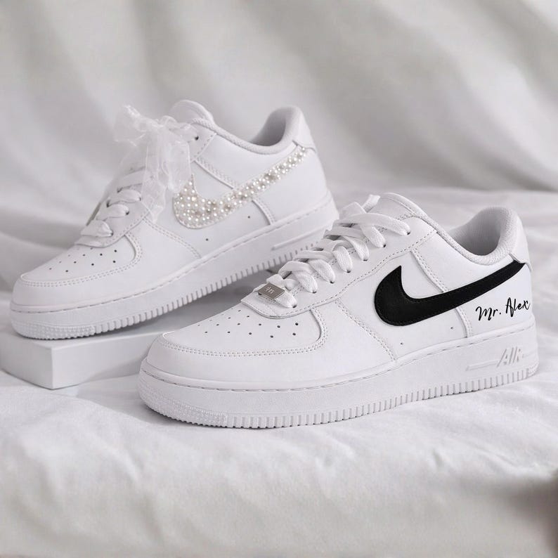 May include: Two white sneakers with black swooshes. One shoe features a pearl embellishment and a sheer ribbon. The other shoe has the text "Mr. Alex" in black script. Both shoes have white laces and soles.