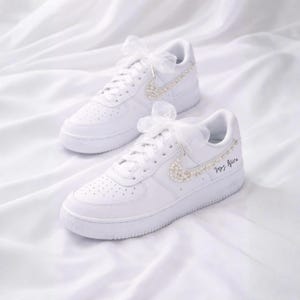 May include: A pair of white sneakers with pearl accents and a sheer bow. The Nike swoosh is embellished with pearls, and the text "Mrs. Alice" is written on the side. The shoes are set against a white fabric background.