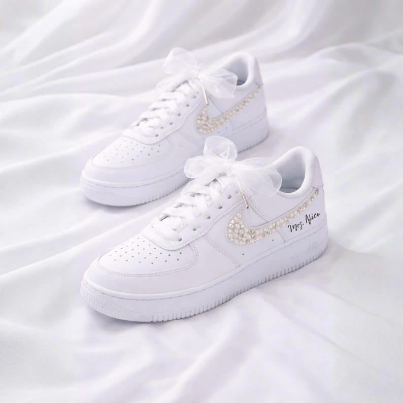 May include: A pair of white sneakers with pearl accents and a sheer bow. The Nike swoosh is embellished with pearls, and the text "Mrs. Alice" is written on the side. The shoes are set against a white fabric background.