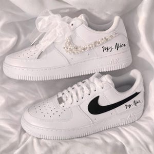 May include: White sneakers with black accents and the text "Mr. Alex" and "Mrs. Alice". One pair features a pearl-studded swoosh and a sheer ribbon bow. The other has a black swoosh. Both have "AIR" text on the sole.