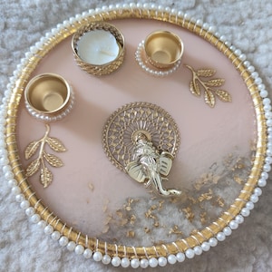 Puede incluir: Una bandeja rosa decorativa con detalles dorados, perlas y una figura de elefante dorada. La bandeja presenta tres pequeños candelabros dorados, una vela y detalles de hojas doradas. La bandeja tiene aproximadamente 25 cm de diámetro.