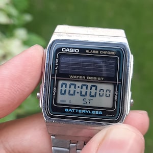 May include: A silver Casio digital watch with a black and teal face. The watch displays "00:0000" and "ST" on the screen. The watch has the text "CASIO ALARM CHRONO" and "WATER RESIST" printed on the face. The watch is held by a person.