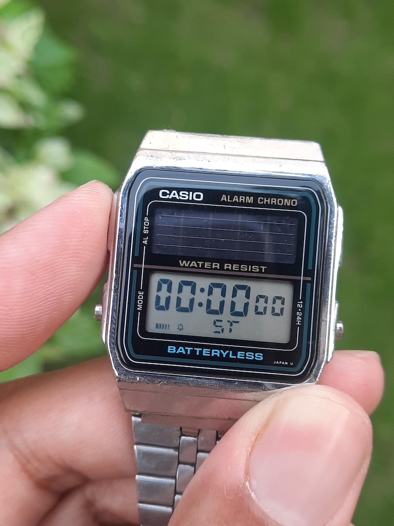 May include: A silver Casio digital watch with a black and teal face. The watch displays "00:0000" and "ST" on the screen. The watch has the text "CASIO ALARM CHRONO" and "WATER RESIST" printed on the face. The watch is held by a person.