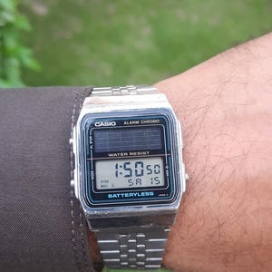 May include: A silver Casio digital watch displaying the time 1:50:50 PM on Saturday the 15th. The watch has a black and teal face with the words "CASIO", "ALARM CHRONO", and "WATER RESIST". The watchband is silver.