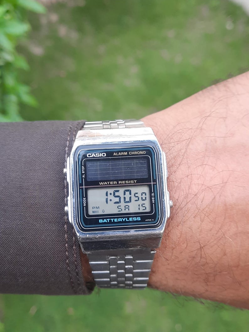 May include: A silver Casio digital watch displaying the time 1:50:50 PM on Saturday the 15th. The watch has a black and teal face with the words "CASIO", "ALARM CHRONO", and "WATER RESIST". The watchband is silver.