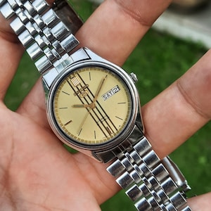 May include: A silver-toned wristwatch with a gold-colored face. The watch face displays the day and date, with the day reading "SAT 21". The watch has a silver band and a classic design.