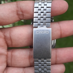 May include: Close-up of a silver-toned Casio watch band. The band is made of small, rectangular metal links. The clasp features the brand name "CASIO" in a stamped font. The watch band is held against a blurred green background.