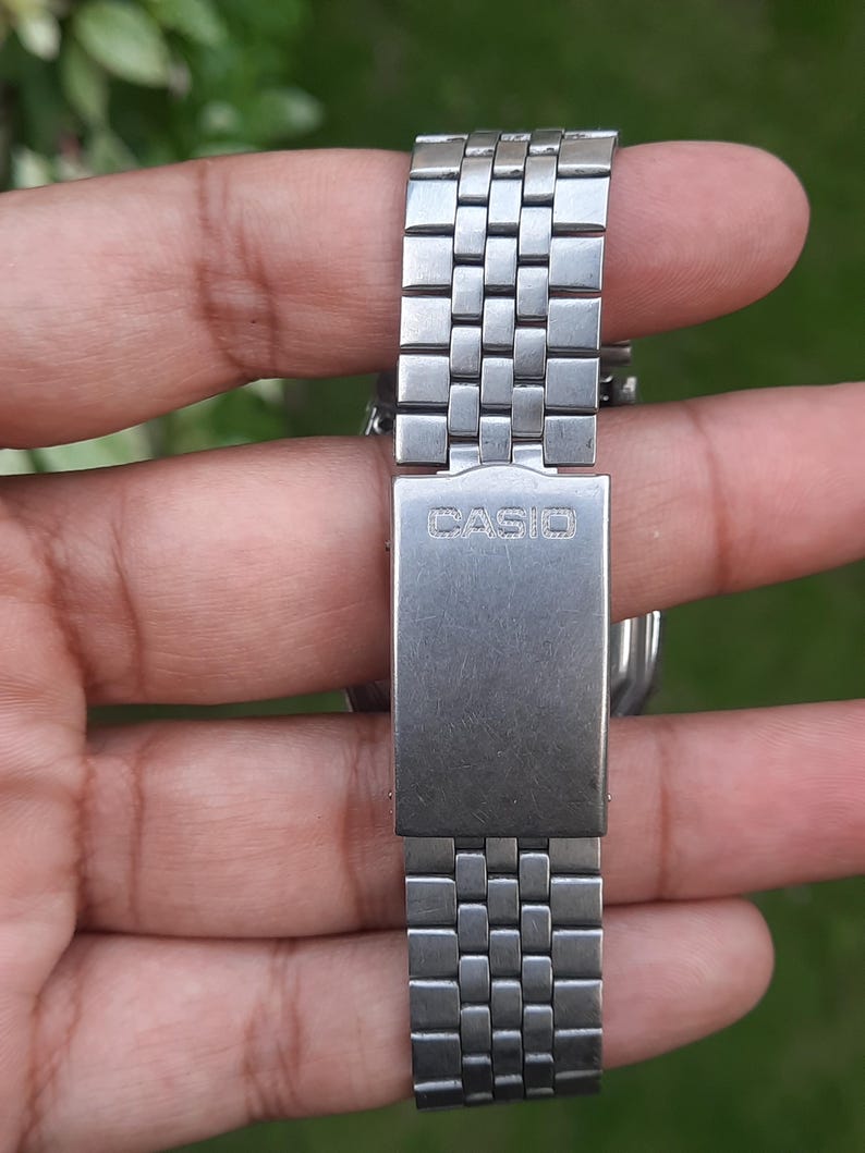 May include: Close-up of a silver-toned Casio watch band. The band is made of small, rectangular metal links. The clasp features the brand name "CASIO" in a stamped font. The watch band is held against a blurred green background.