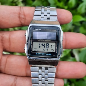May include: A silver Casio digital watch with a rectangular face and a stainless steel band. The watch displays the time, date, and other functions on a black screen. The watch is labeled "WATER RESIST" and "BATTERYLESS".
