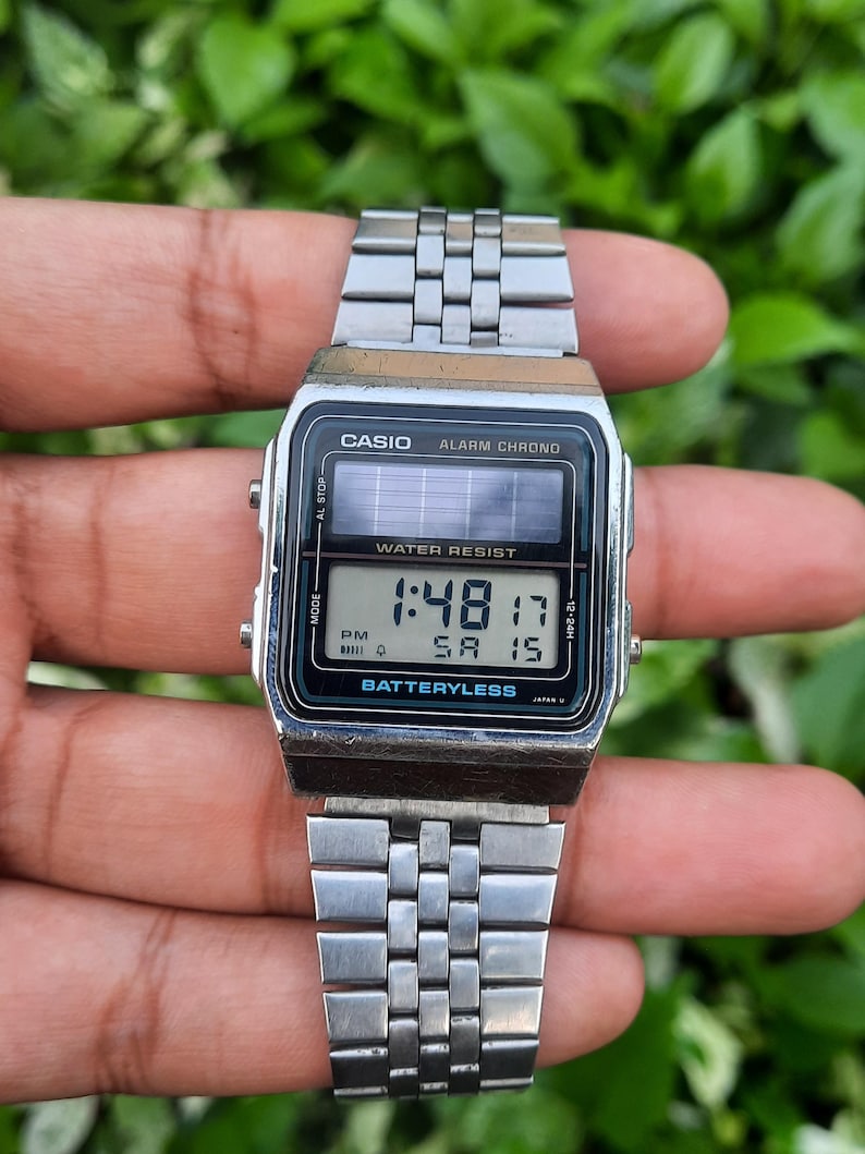 May include: A silver Casio digital watch with a rectangular face and a stainless steel band. The watch displays the time, date, and other functions on a black screen. The watch is labeled "WATER RESIST" and "BATTERYLESS".