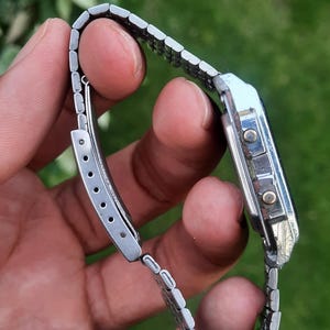 May include: A silver-toned digital wristwatch with a rectangular face and a linked metal band. The watch has two buttons on the side and a clasp closure. The watch is shown against a blurred green background.