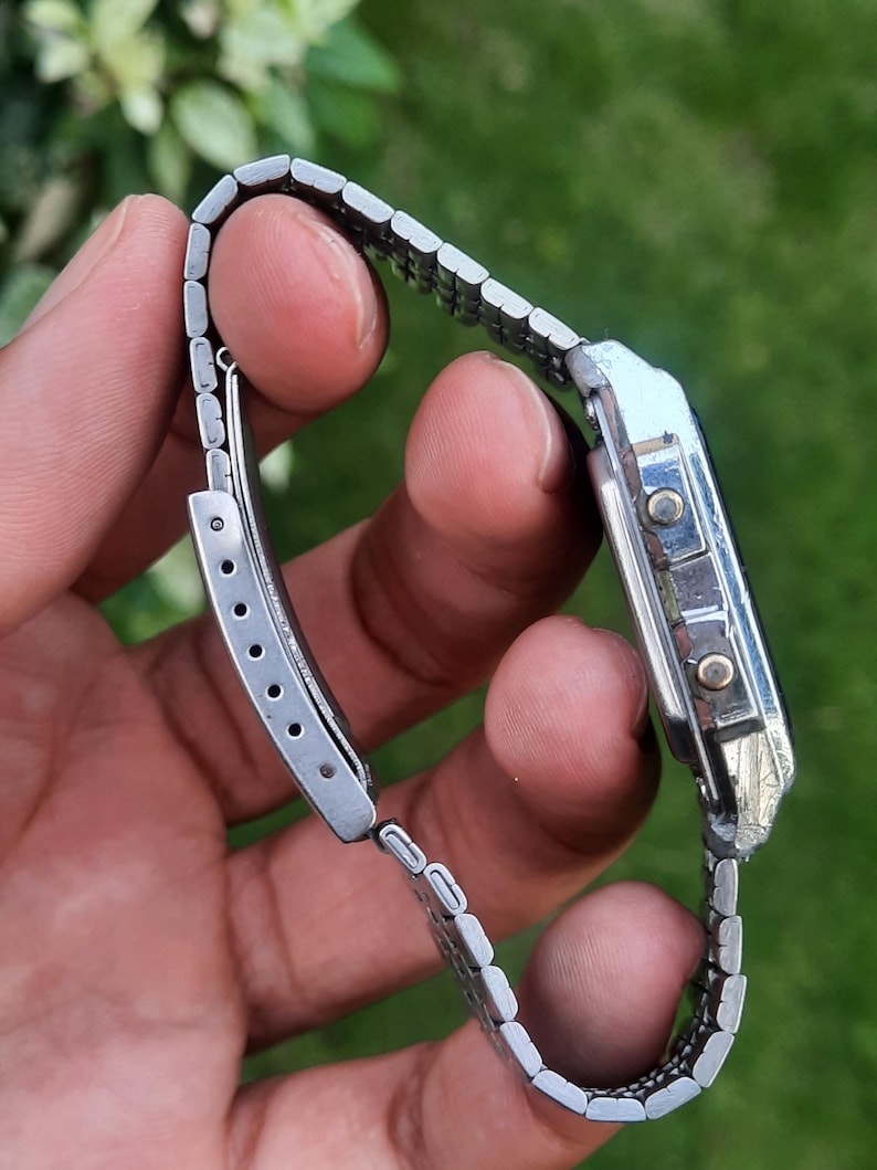 May include: A silver-toned digital wristwatch with a rectangular face and a linked metal band. The watch has two buttons on the side and a clasp closure. The watch is shown against a blurred green background.