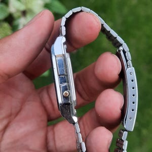 May include: A silver-toned digital watch with a rectangular face and a linked metal band. The watch has a small gold-colored button and a black display. The band is adjustable and has a clasp closure.