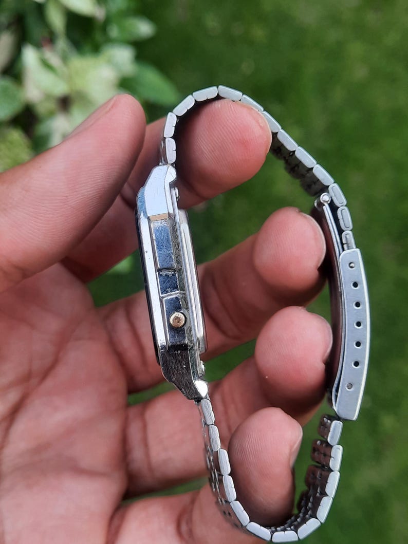 May include: A silver-toned digital watch with a rectangular face and a linked metal band. The watch has a small gold-colored button and a black display. The band is adjustable and has a clasp closure.