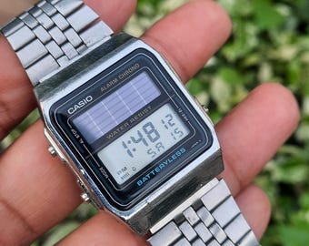 Puede incluir: Un reloj digital Casio plateado con una esfera rectangular y una correa de metal. El reloj muestra la hora 1:48 PM un sábado. La esfera del reloj tiene las palabras "CASIO", "ALARM CHRONO", "WATER RESIST" y "BATTERYLESS".